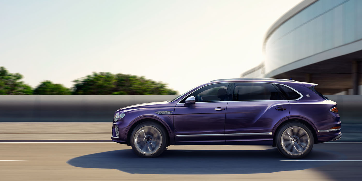 Bentley Dubai Bentley Bentayga Extended Wheelbase Mulliner luxury SUV profile in Tanzanite Purple paint, driving on an country road past trees.