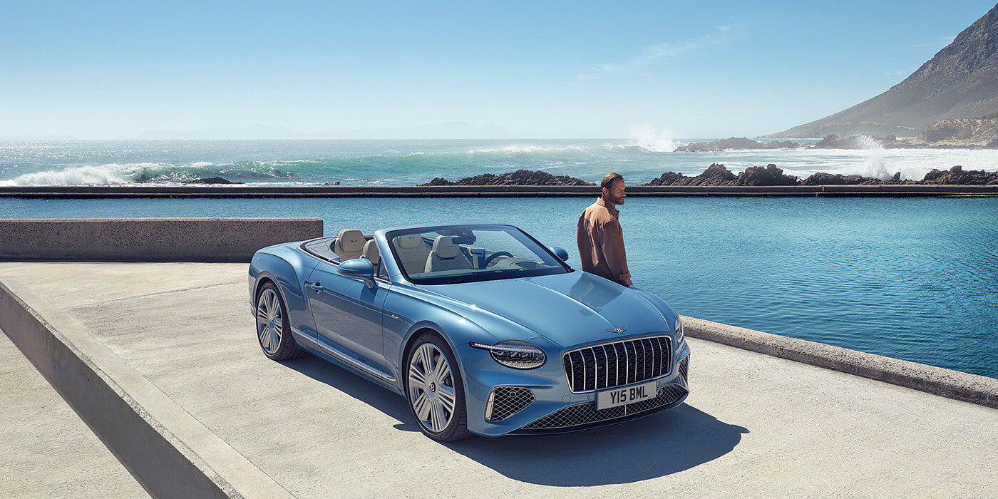 Bentley Dubai New Bentley Continental GTC Azure convertible front view in Light Windsor blue paint with 22 inch ten spoke wheels parked by the sea in the sunshine
