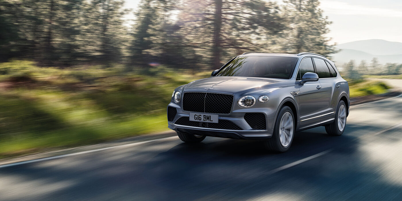 Bentley Dubai Bentley Bentayga luxury SUV front three quarter in Moonbeam paint, driving on an country road past trees.
