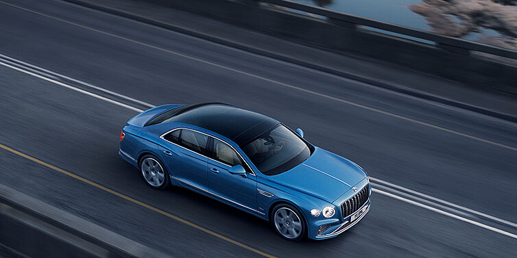 Bentley Dubai New Bentley Flying Spur Azure sedan in Light windsor blue paint by Mulliner, with 22 inch Azure wheels seen from overhead driving dynamically on a country road