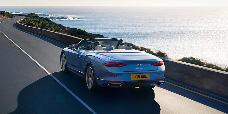 Bentley Dubai New Bentley Continental GTC Azure convertible rear view in Light Windsor blue paint with 22 inch ten spoke wheels driving dynamically on an ocean road in the sunshine