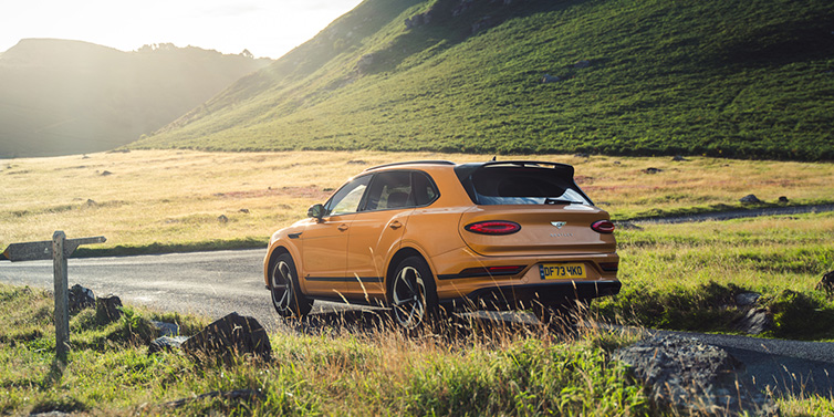 Bentley Dubai Bentley Bentayga S SUV rear three quarter in Sunburst Gold paint on a road surrounded by green hills