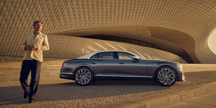 Bentley Dubai Bentley Flying Spur Mulliner sedan in Rose Gold over Magnetic duotone paint parked by a curved wall with person in suit jacket standing on the left